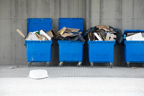Six blue wheelie bins lined up on a paved street surface in an urban area, with three of them filled with various types of rubbish including cardboard boxes, paper debris, and black plastic bags, some of which are partially open, revealing their contents. The bins are positioned against a plain concrete wall and are likely part of a waste collection routine, possibly in a residential or commercial setting in Spitalfields, with some waste spilling slightly over the edges. The scene captures typical rubbish disposal with different materials stacked within the bins, emphasizing the importance of waste management services such as those offered by Garden Clearance Spitalfields. A white discarded paper or cardboard item lies on the ground in front of the bins. The environment is clean with a neutral lighting condition, highlighting the textures and colors of the waste and the bins, emphasizing an everyday urban waste disposal scenario relevant to rubbish removal needs in the area.