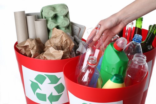 Close-up image of two red recycling bins placed outdoors on a paved surface, filled with assorted waste materials. The left bin contains rolled-up brown and grey cardboard tubes, crumpled brown paper, and an egg carton made of green pulp. The right bin is filled with clear plastic bottles, some with labels or caps, and a few green glass bottles, all arranged with some standing upright and others leaning. The background is out of focus, emphasizing the contents of the bins. This scene illustrates waste segregation for recycling, typical of rubbish removal services in urban areas like Spitalfields. The bins feature a green recycling symbol on their white labels, indicating proper waste separation. The lighting appears natural, with a soft, even illumination highlighting textures such as the glossy plastic bottles, the matte cardboard, and the crumpled paper, emphasizing the diversity of materials requiring collection and disposal as part of professional rubbish removal in London neighborhoods.