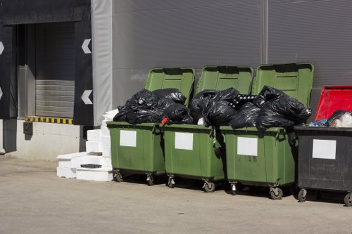 Four large wheeled rubbish bins are positioned on a paved surface next to a building with gray exterior walls. The first three bins are green with open lids, filled with tightly packed black rubbish bags. The fourth bin, also green, appears to be partially open, revealing more black waste bags inside. To the right of these bins, there is a single red bin, also with its lid partially open, containing additional rubbish. Behind the bins, a slow-moving industrial roller shutter door is visible, with a yellow and black striped safety barrier marking its edge. The surroundings suggest an urban or commercial area, possibly a back alley or service entrance, consistent with waste collection and rubbish removal services in Spitalfields, London. Garden Clearance Spitalfields manages waste collection for properties in this postcode area, providing professional rubbish disposal solutions.