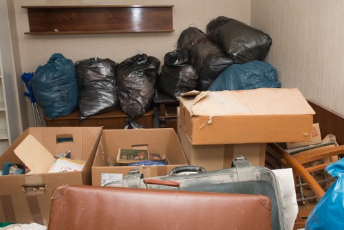 The image displays a cluttered indoor space with multiple black and blue garbage bags stacked against a beige wall, some leaning on a wooden shelf. In the foreground, there are open cardboard boxes filled with various rubbish items, including paper and small household objects. A large metal vacuum cleaner or similar appliance lies partially visible at the bottom right corner, with a brown leather or faux-leather piece, possibly a cushion or furniture padding, placed in front of it. To the right, leaning on or partly covering the boxes, is a wooden laundry drying rack. The environment appears to be a storage or utility area, with minimal natural light. This setting suggests a collection of waste or discarded items ready for rubbish removal, consistent with services offered by Garden Clearance Spitalfields, serving the E1 postal district in London.