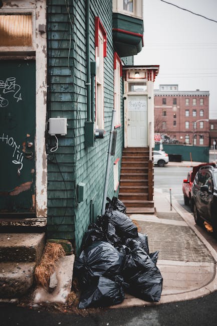 An urban street scene with multi-story buildings showcasing a mix of architectural styles, including brick facades, large bay windows, and ornate details. The foreground features a white waste collect